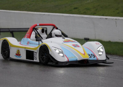 Team Earle’s #39 Radical SR3 XX, painted white, blue, and yellow, driving on a race track slick with water due to rainfall
