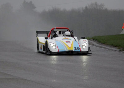 Team Earle’s #39 Radical SR3 XX, painted white, blue, and yellow, driving on race track slick with water due to rainfall, with another race car and a technician in safety fatigues partially visible in the background