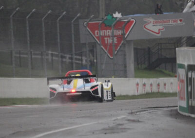 Team Earle’s #39 Radical SR3 XX, painted white, blue, and yellow, visible from the rear as it speeds down a race track underneath a viewing station displaying the Canadian Tire logo