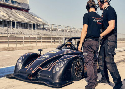 Team Earle’s #39 Radical SR3 XX, painted black making a pit stop on a motor speedway with audience seating in the background