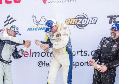 Daniel Earle, captain of Team Earle and president and CEO of Highlander Silver, on a podium popping the cork off a bottle of Champagne, joined by two other drivers