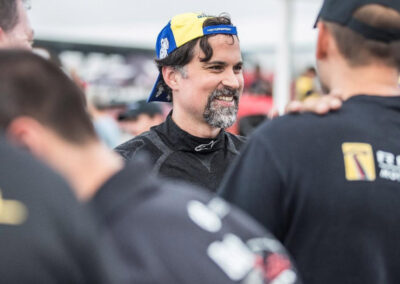Daniel Earle, captain of Team Earle and president and CEO of Highlander Silver, wearing racing fatigues and a backwards baseball cap, speaking with other racers