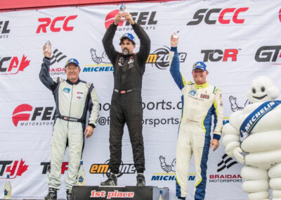 Daniel Earle, captain of Team Earle and president and CEO of Highlander Silver, atop a podium raising a trophy over his head accompanied by two other drivers and the Michelin mascot