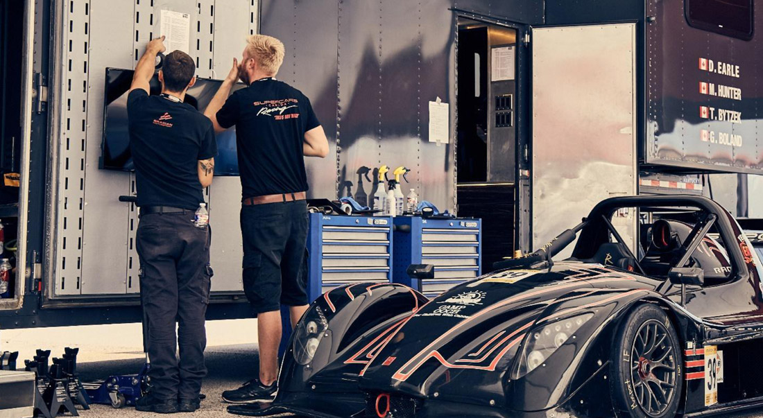 home-img Two race team members examining a document taped to a trailer next to Team Earle’s #39 Radical SR3 XX, painted black